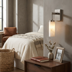 Mounted beside a bed's nightstand, this brushed-nickel Clérachic sconce casts a warm glow over a woven basket and books.