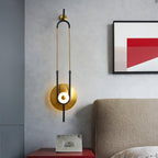 A Clérachic gold wall sconce glows above a red-lacquered nightstand, its black-and-gold loop rising past a framed abstract print in a bedroom.