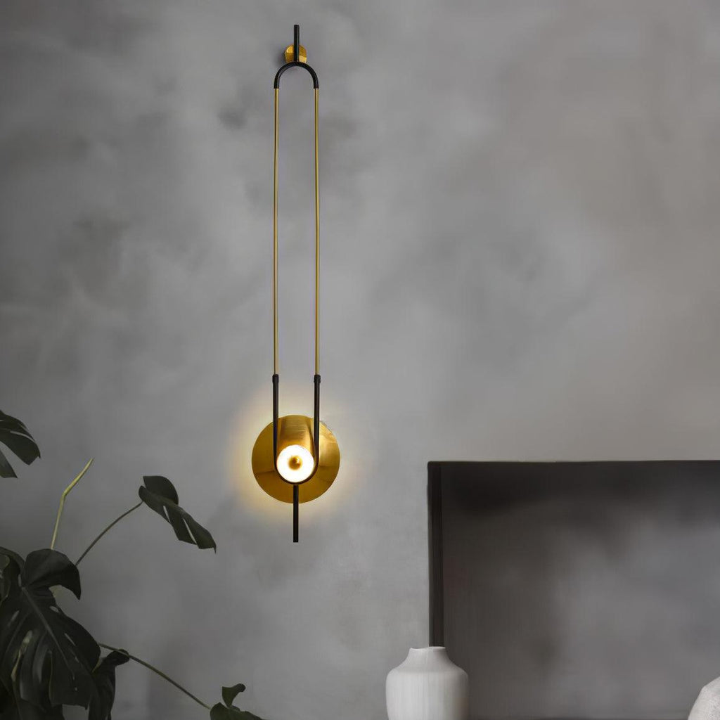 A Clérachic wall sconce lights a textured grey bedroom wall above a potted plant, its gold disc glowing beside the headboard.