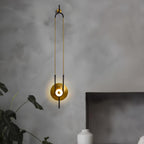 A Clérachic wall sconce lights a textured grey bedroom wall above a potted plant, its gold disc glowing beside the headboard.