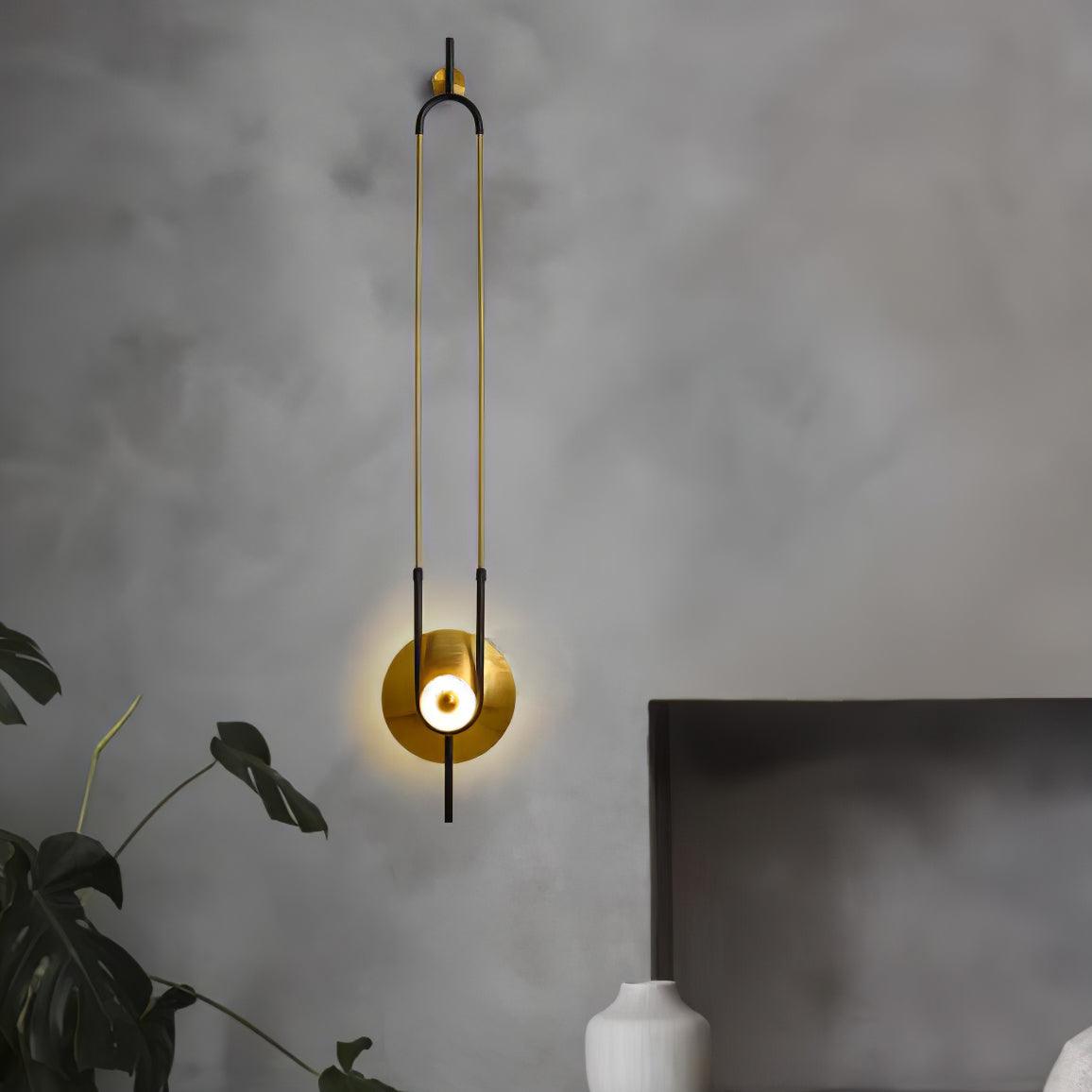 A Clérachic wall sconce lights a textured grey bedroom wall above a potted plant, its gold disc glowing beside the headboard.