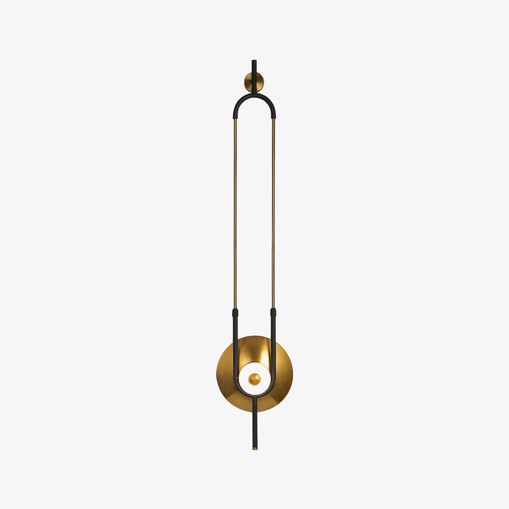 A slim brass loop frame on this Clérachic wall sconce curves down to a round gold disc and a softly lit glass orb, shown alone on a plain backdrop.