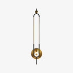 A slim brass loop frame on this Clérachic wall sconce curves down to a round gold disc and a softly lit glass orb, shown alone on a plain backdrop.