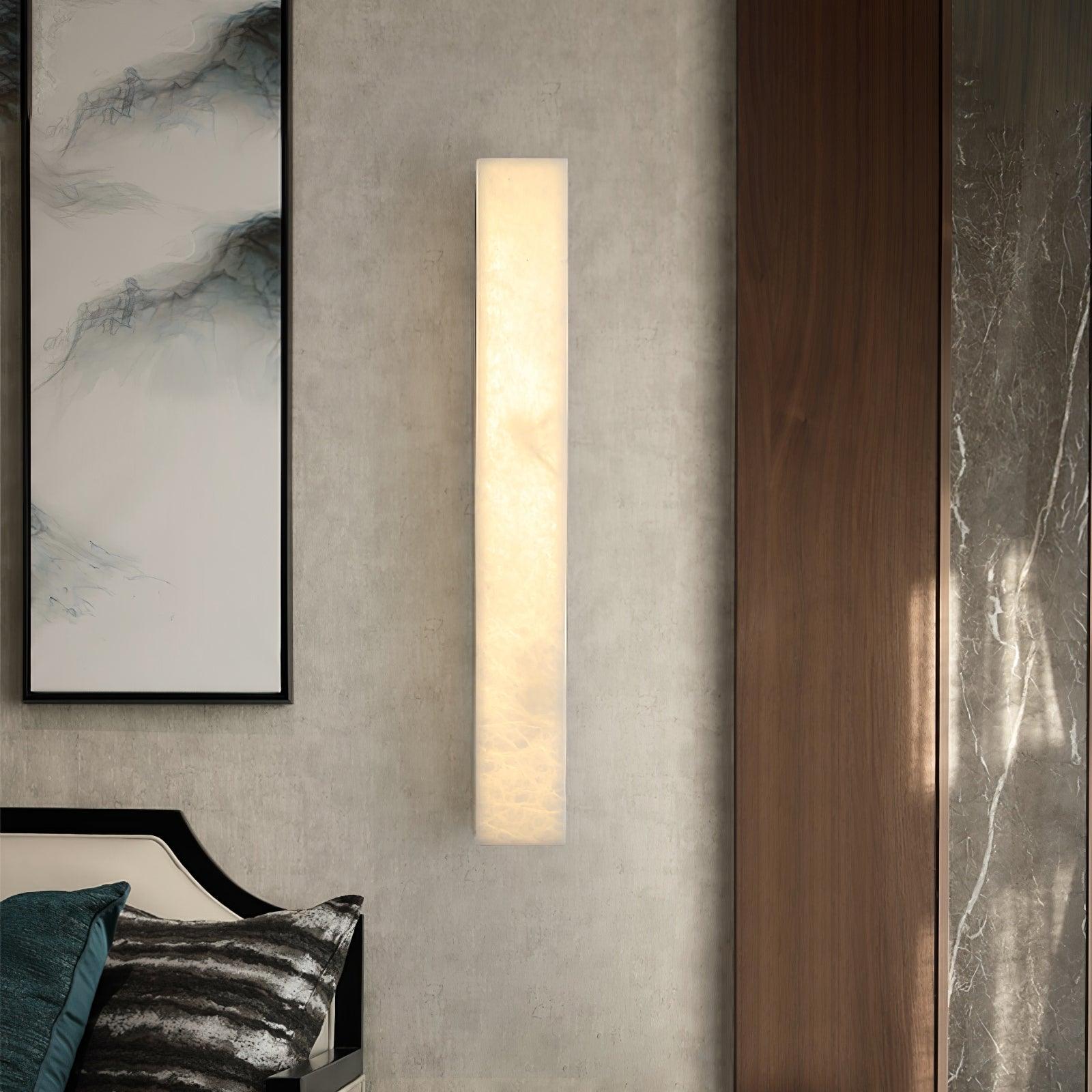 Kristy alabaster wall lamp glowing on a concrete-toned bedroom wall beside walnut and marble.