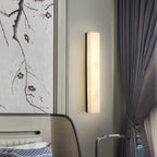 Black-framed Kristy wall lamp lit beside a bed, next to a blossom-branch panel and grey drapes.