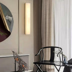 Kristy wall lamp lit on a cream panelled wall beside a round gilt-framed painting and dark chair.