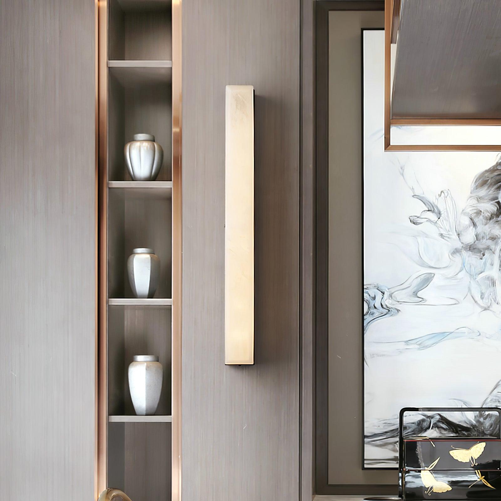 Unlit Kristy wall lamp beside a display niche of silver vases on a grey-brown panelled wall.