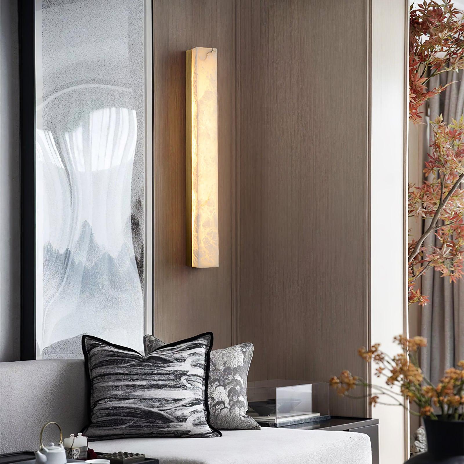 Brass Kristy wall lamp lit on a timber panelled wall above a sofa with marbled cushions.