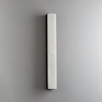 Unlit black-framed Kristy wall lamp on a grey wall, cool white stone face and dark edge.
