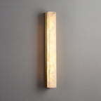 Slim brass Kristy wall lamp lit on a grey wall, warm light spilling along the stone face.