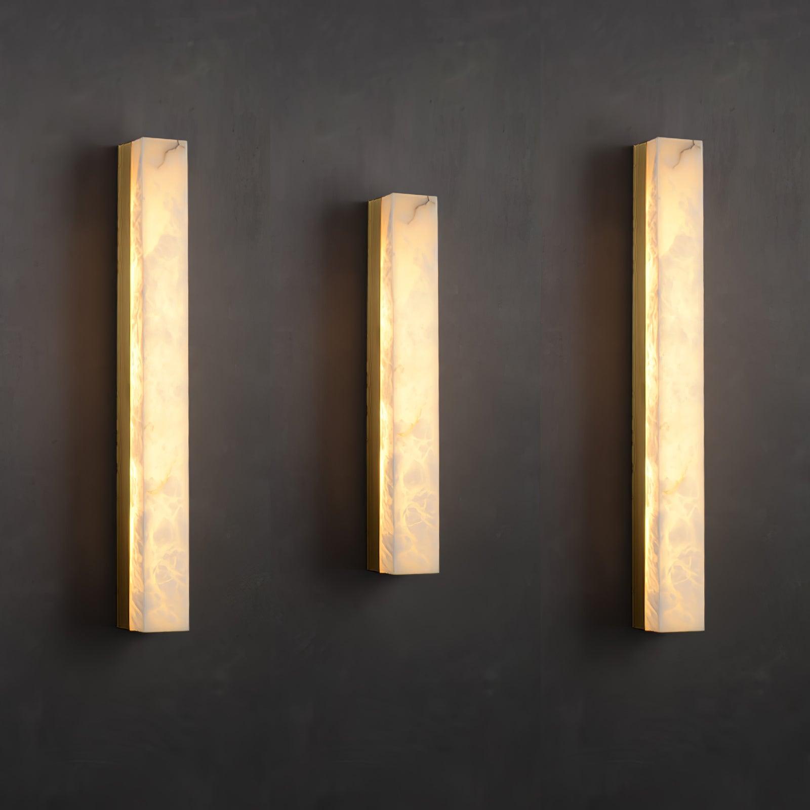 Three lit Kristy wall lamps in different heights mounted side by side on a dark charcoal wall.