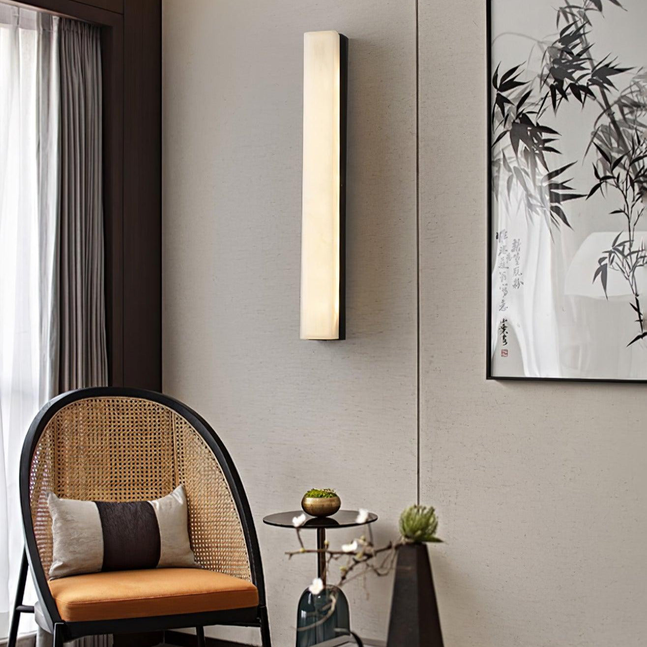 Kristy wall lamp lit in a corner above a cane armchair, beside a bamboo ink painting.