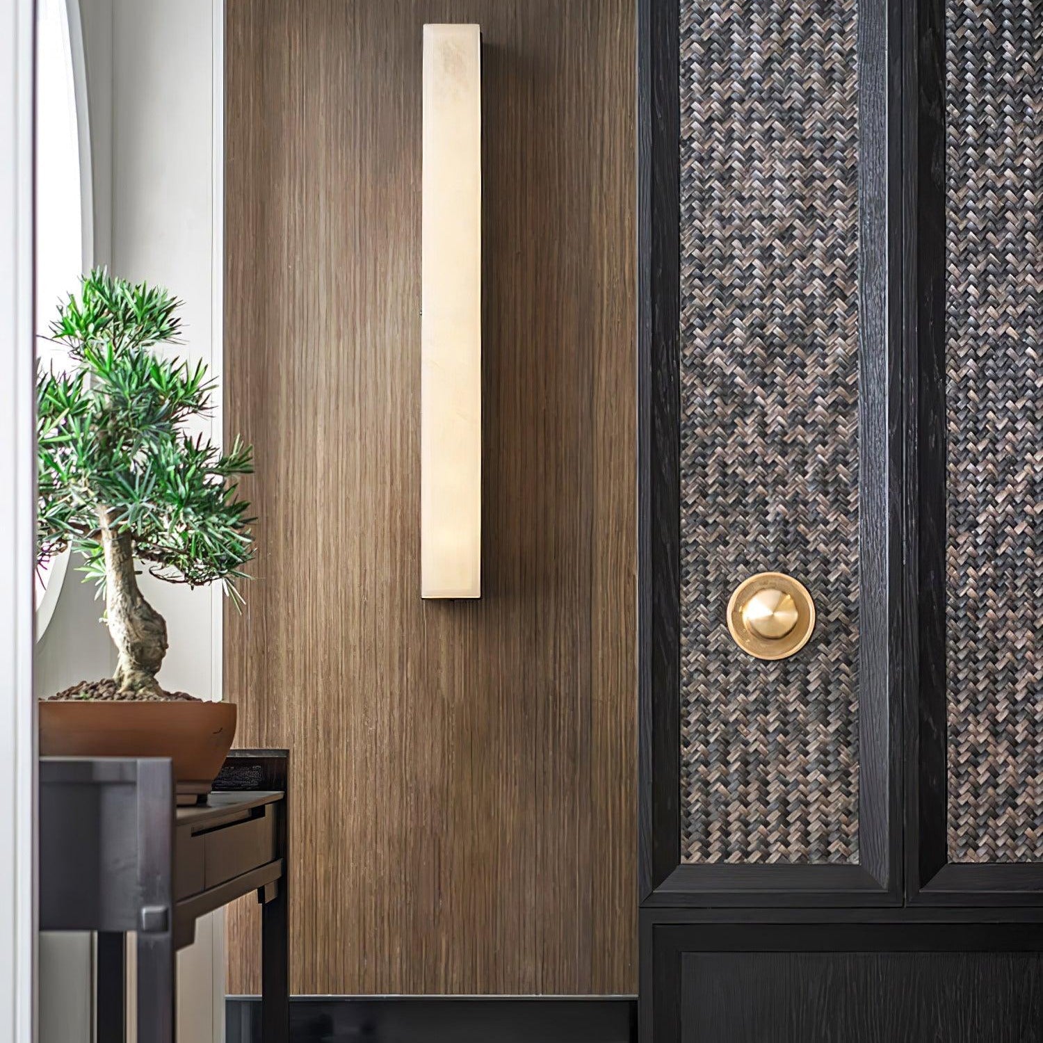 Kristy alabaster wall lamp lit on a walnut panelled entry wall beside a woven cabinet door.
