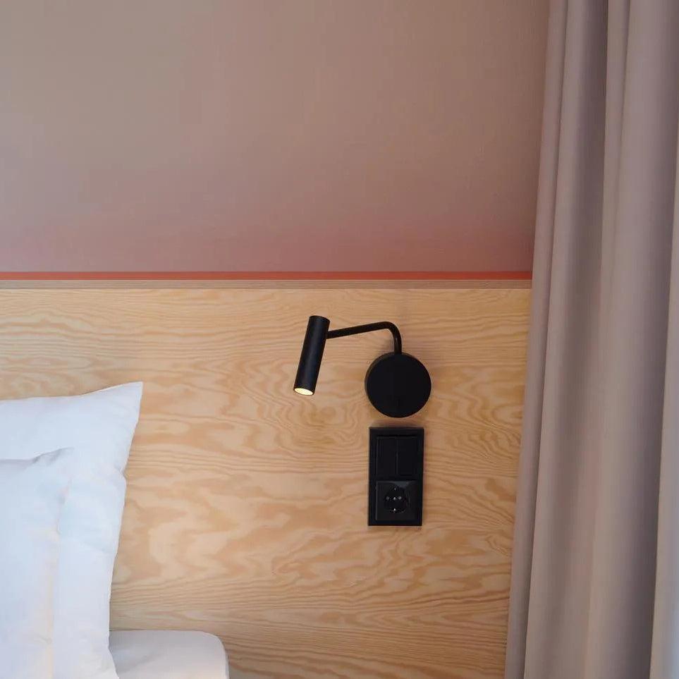 A black Clérachic wall sconce fixed to a wood headboard beside a pink wall and a socket.