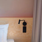 A black Clérachic wall sconce fixed to a wood headboard beside a pink wall and a socket.