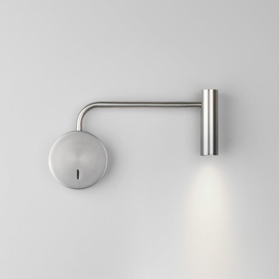 A sand nickel Clérachic wall sconce with a long arm casts light down a plain white wall.