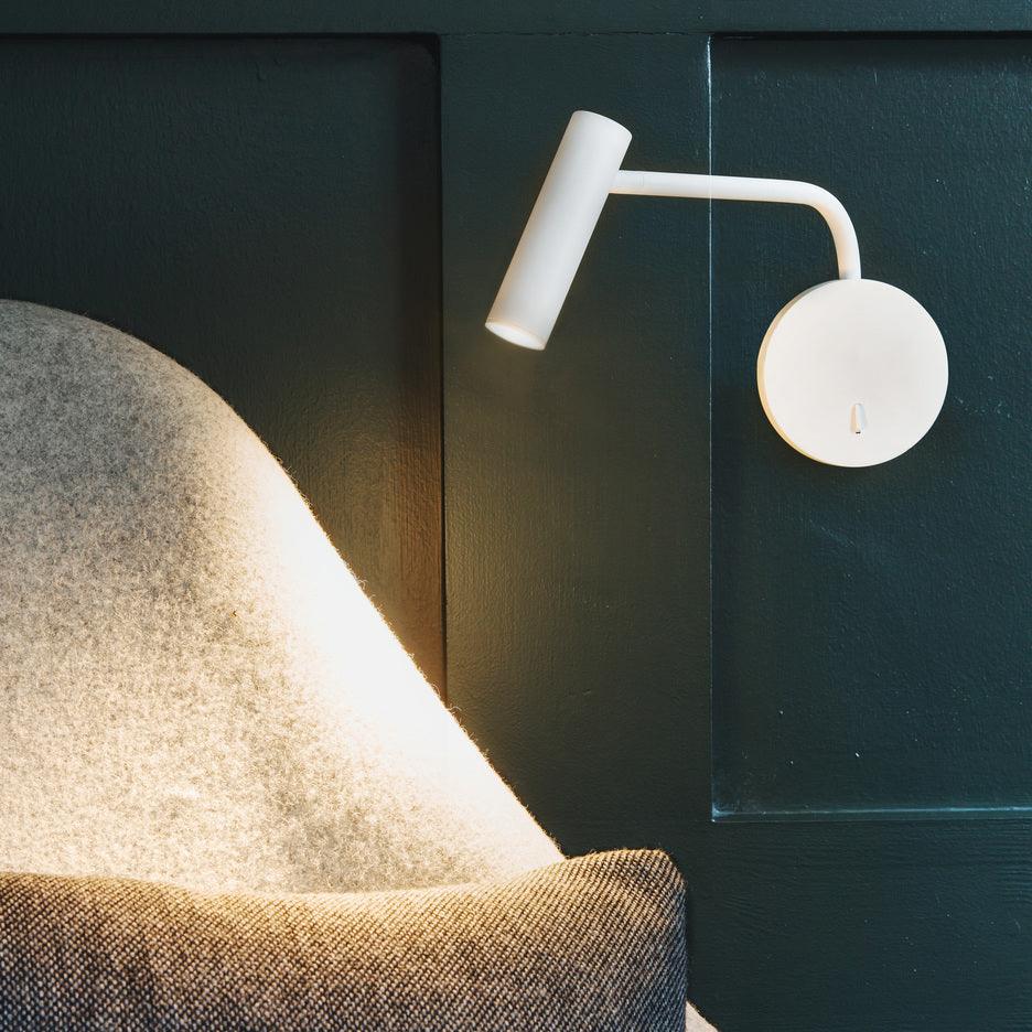 A white Clérachic wall sconce glows above a tufted armchair on a dark teal panelled wall.