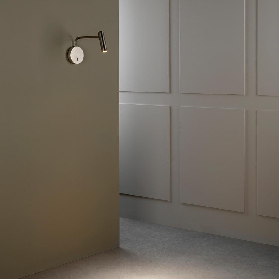 A sand nickel Clérachic wall sconce lit in a dim hallway with panelled walls.