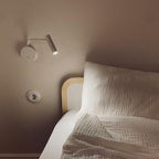 A white Clérachic wall sconce mounted above a bed with a textured pillow and a wall outlet nearby.