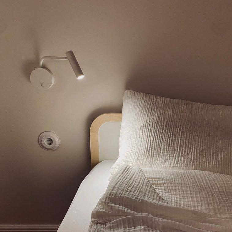 A white Clérachic wall sconce mounted above a bed with a textured pillow and a wall outlet nearby.