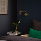 A gold Clérachic wall sconce lit above a marble side table in a dark navy bedroom corner.
