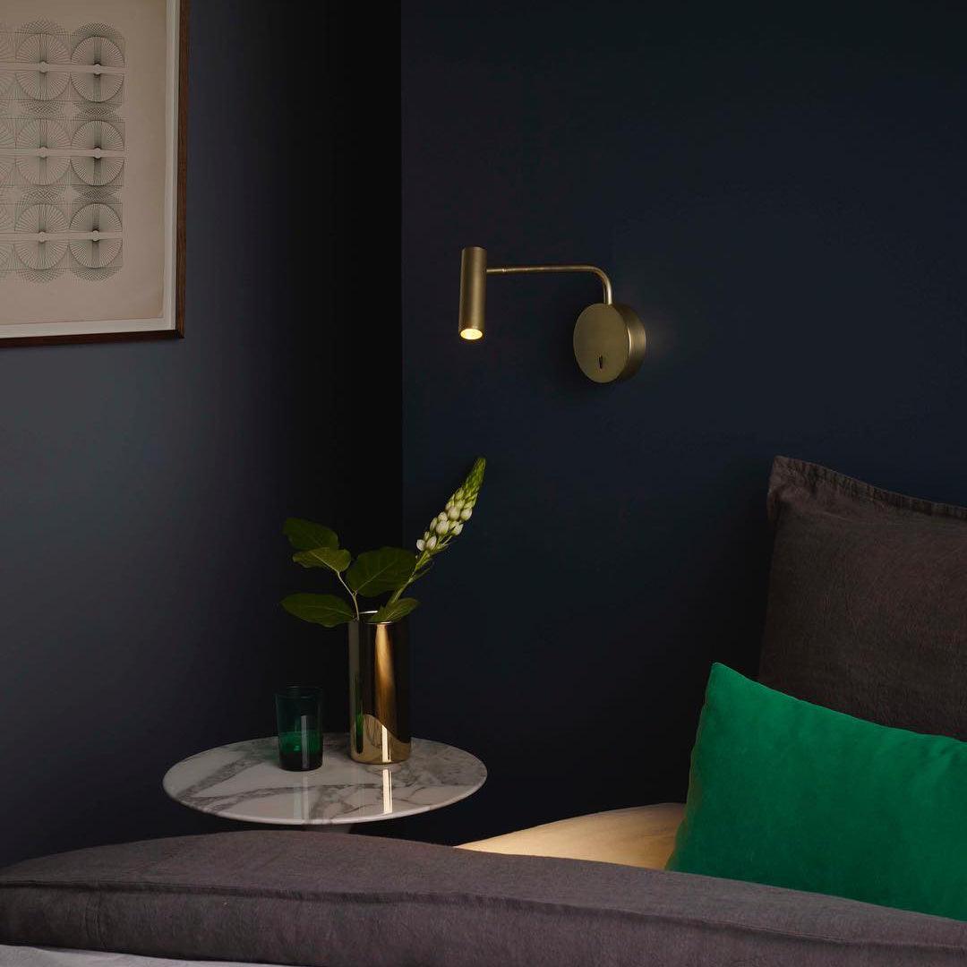 A gold Clérachic wall sconce lit above a marble side table in a dark navy bedroom corner.