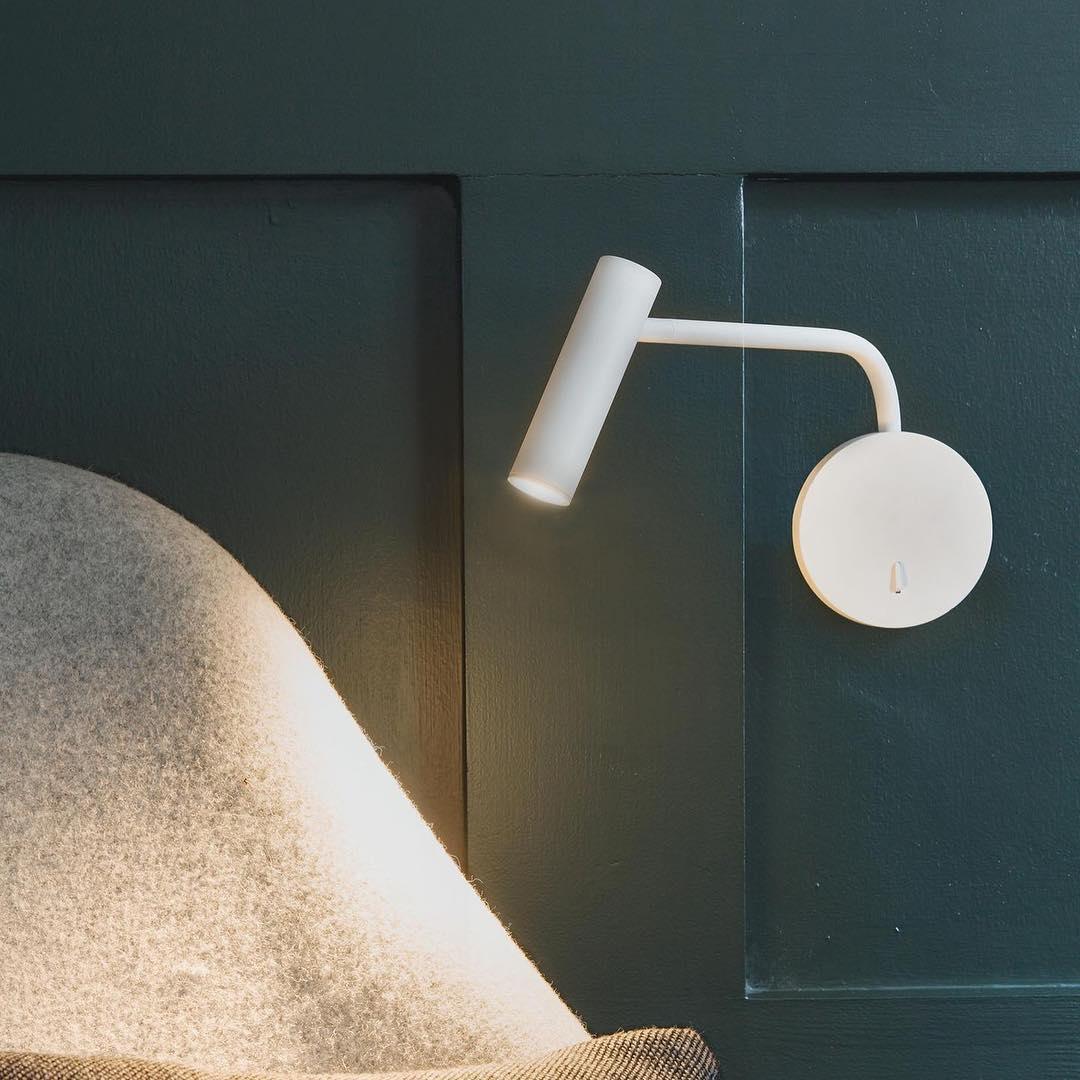 A white Clérachic wall sconce glows above a pillow on a dark teal panelled wall.