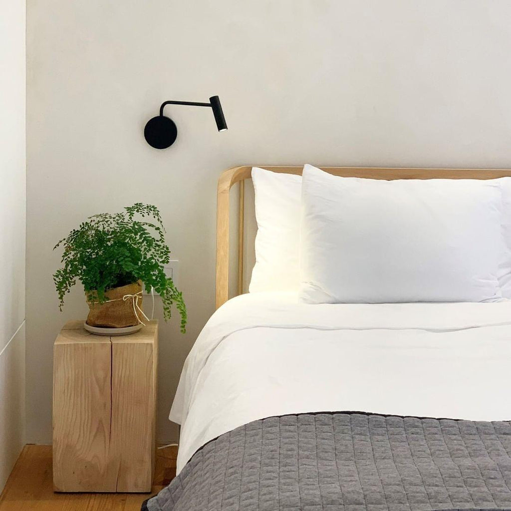 A black wall sconce from Clérachic mounted above a bed with a wood nightstand and a potted plant.