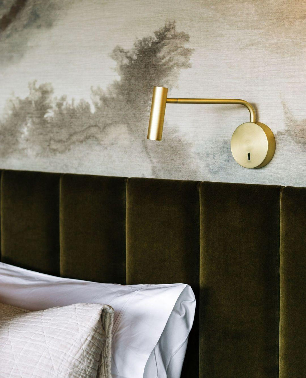 A gold Clérachic wall sconce mounted above a moss-green velvet headboard with a white pillow.