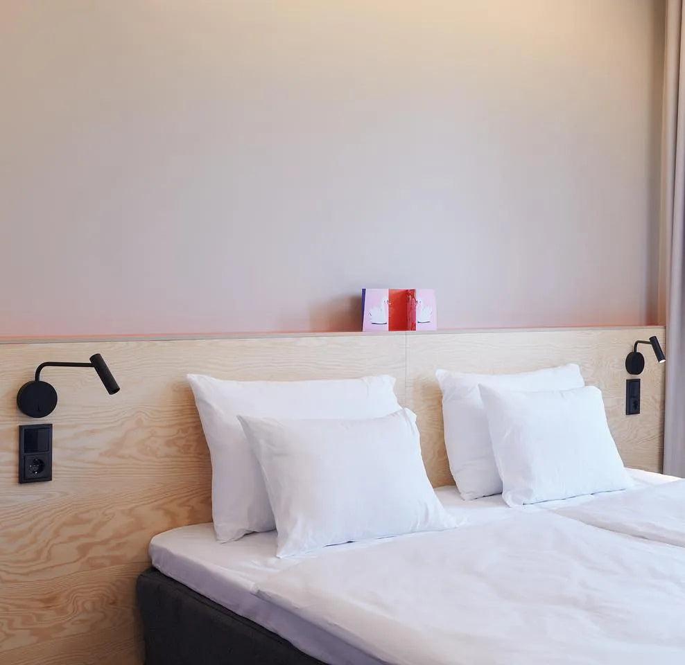 Two black Clérachic wall sconces flank a bed with white pillows on a light wood headboard.