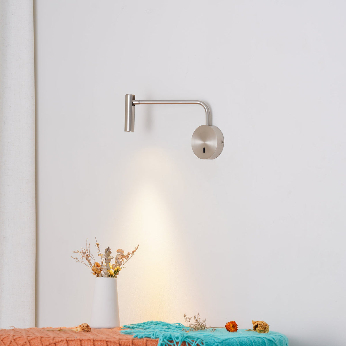 Above a bench with dried flowers and a teal throw, this sand nickel Clérachic wall sconce glows warmly.