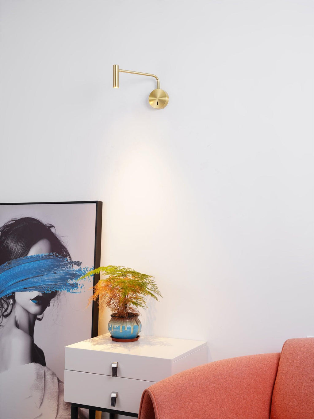 A gold Clérachic wall sconce glows above a white bedside dresser with a small potted plant.