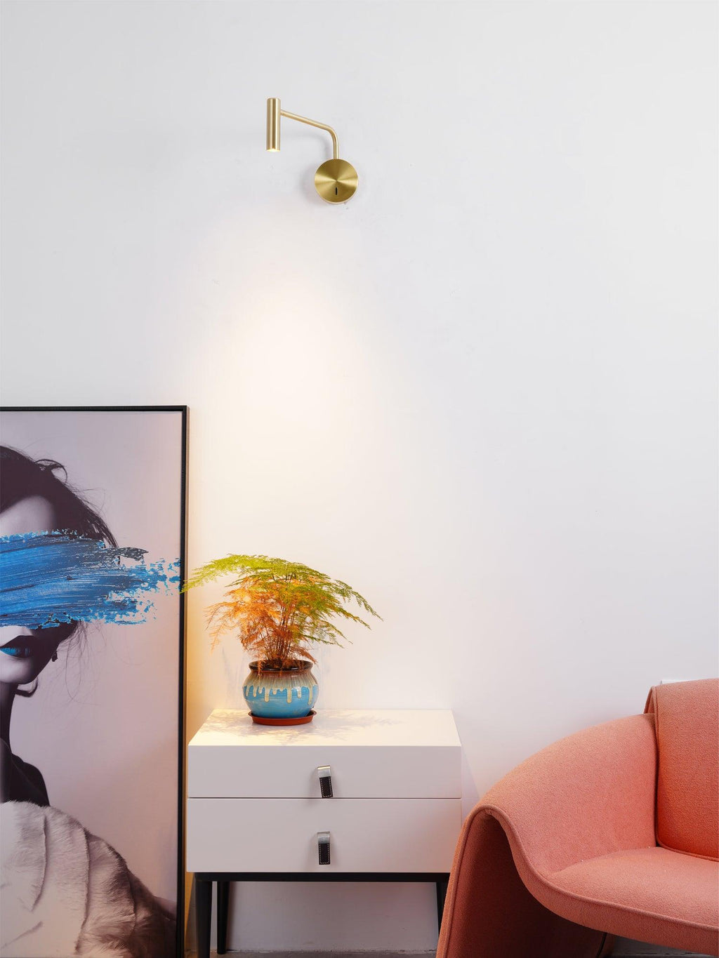A gold Clérachic wall sconce glows above a white dresser with a plant and framed portrait art.