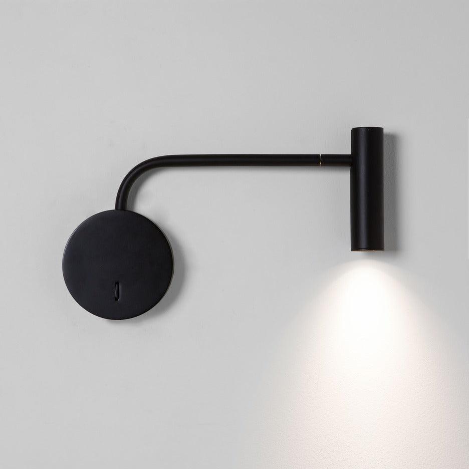 A black Clérachic wall sconce with a long arm casts a warm downward beam on a white wall.