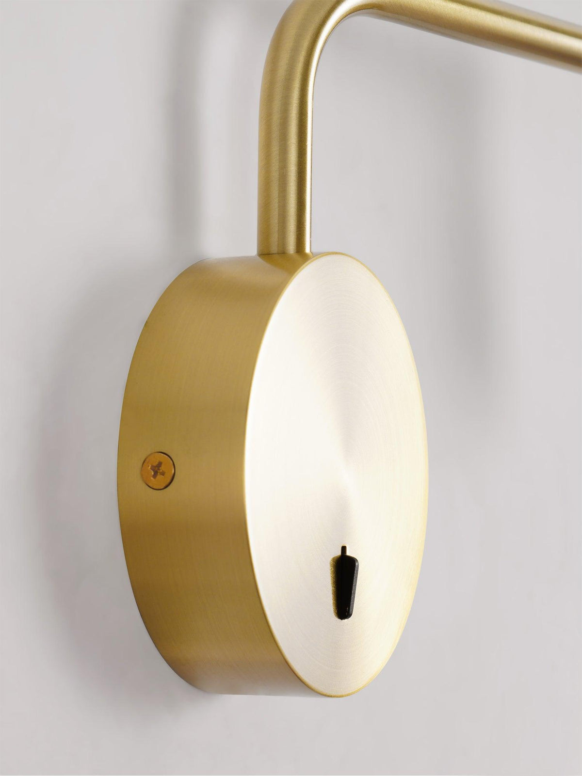 Close view of the gold disc base and curved arm on this Clérachic wall sconce, against a plain backdrop.