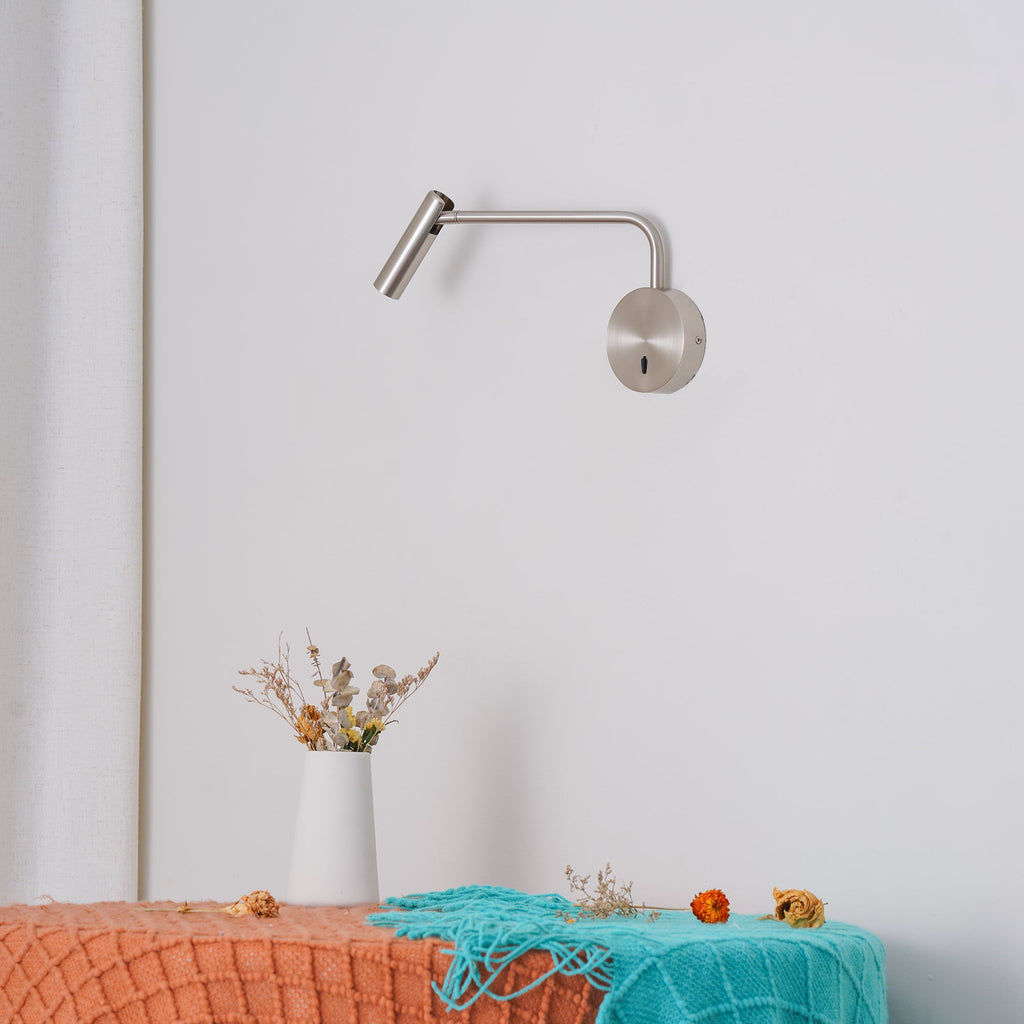 A sand nickel Clérachic wall sconce reaches out from a white wall above a bench with dried flowers.