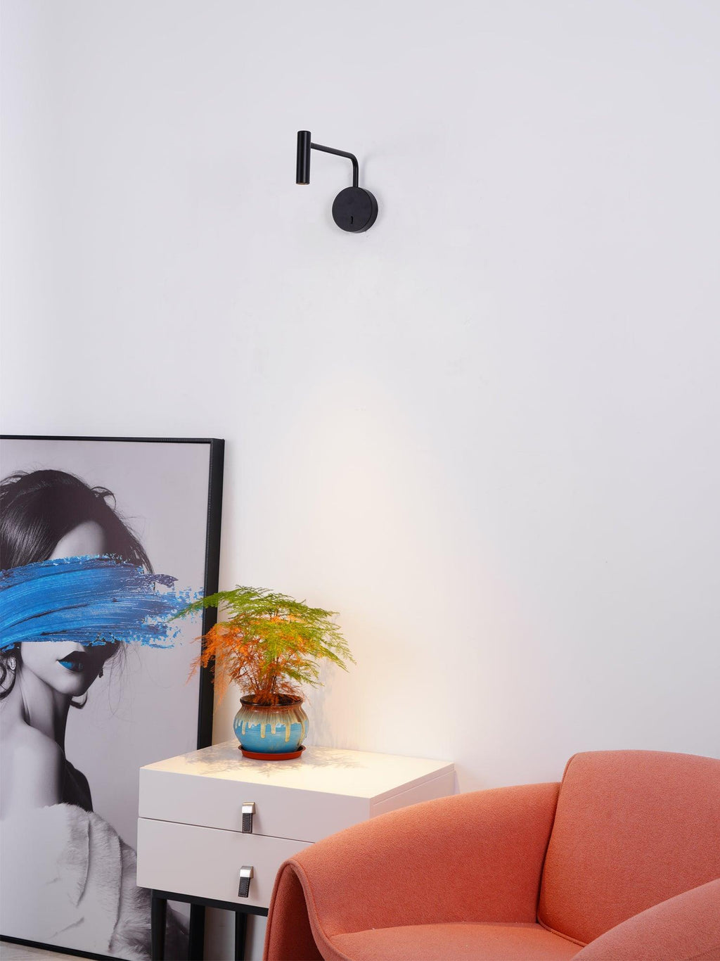 A black Clérachic wall sconce glows above a white dresser beside a coral armchair and framed art.