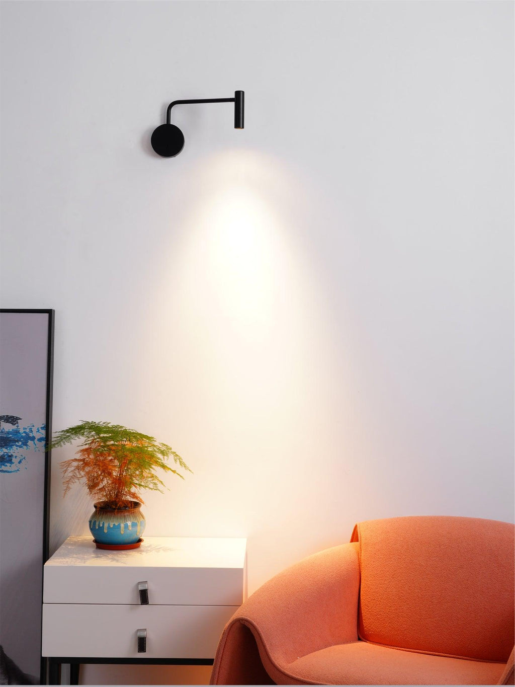 A black Clérachic wall sconce lit above a white dresser beside a coral armchair.