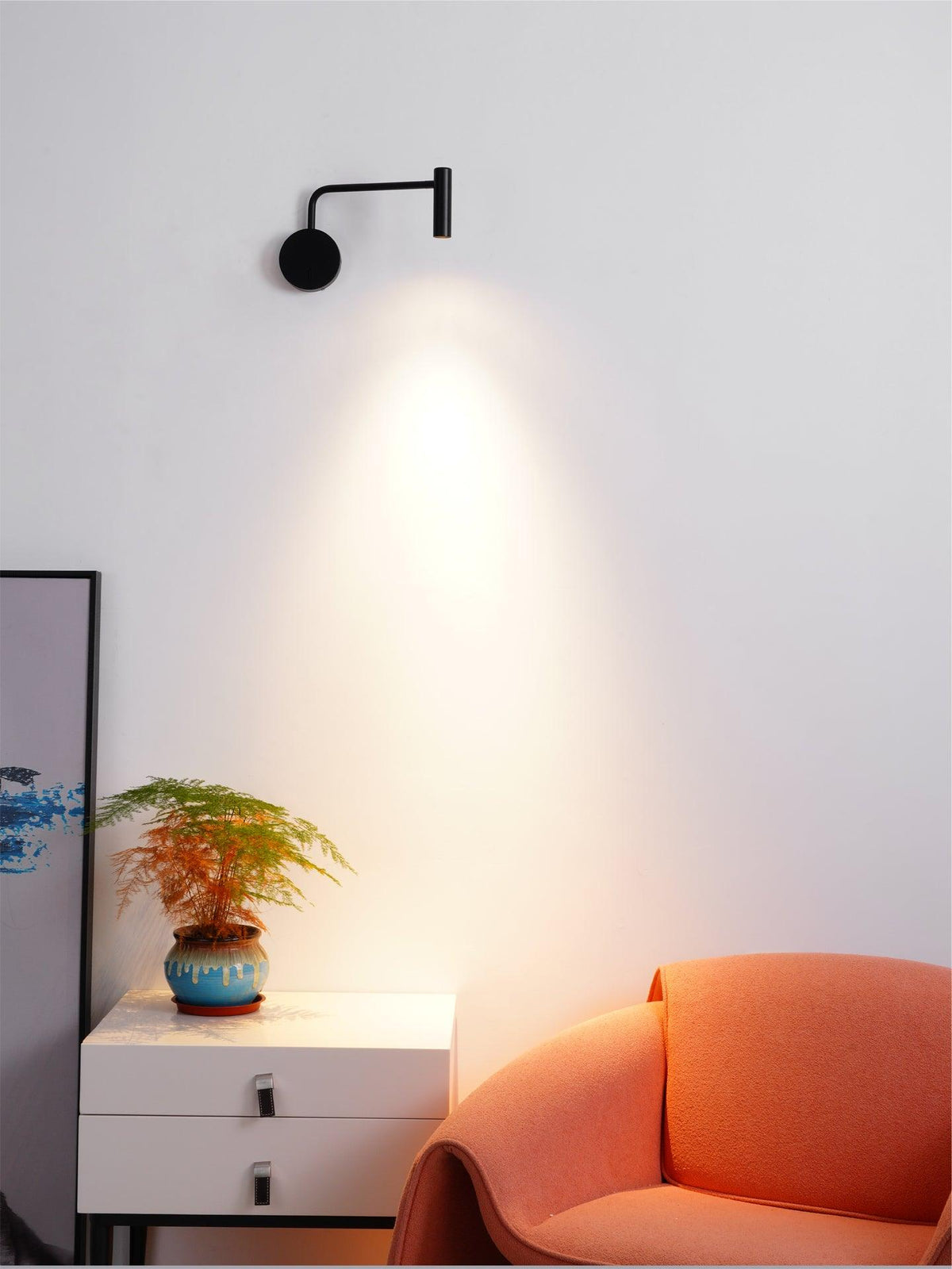 A black Clérachic wall sconce lit above a white dresser beside a coral armchair.