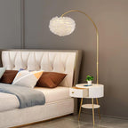 Lit warmly beside the bed, the Clérachic feather shade glows gold against a beige tufted headboard, with a plant on the marble shelf below.