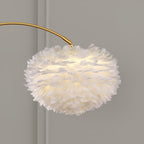 Close-up of the Clérachic feather shade lit from within, showing the layered white plumes against a plain grey wall behind the gold arc.