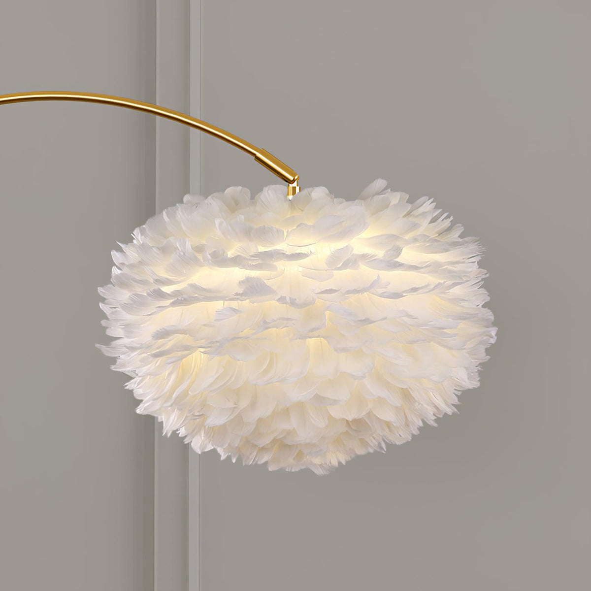 Close-up of the Clérachic feather shade lit from within, showing the layered white plumes against a plain grey wall behind the gold arc.