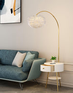 In a living room corner, this Clérachic gold arc lamp arches its feather shade over a teal sofa, the marble side table tucked beside it.