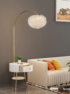 The same Clérachic gold arc floor lamp curves over a cream sofa, its round marble tabletop and gold tripod legs set on a patterned rug.