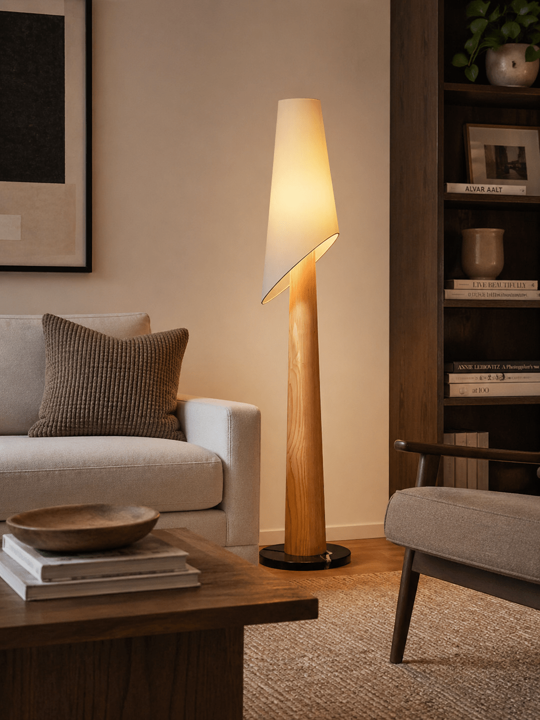 A lit Clérachic floor lamp stands beside a cream sofa in a living room, near a low table stacked with books.