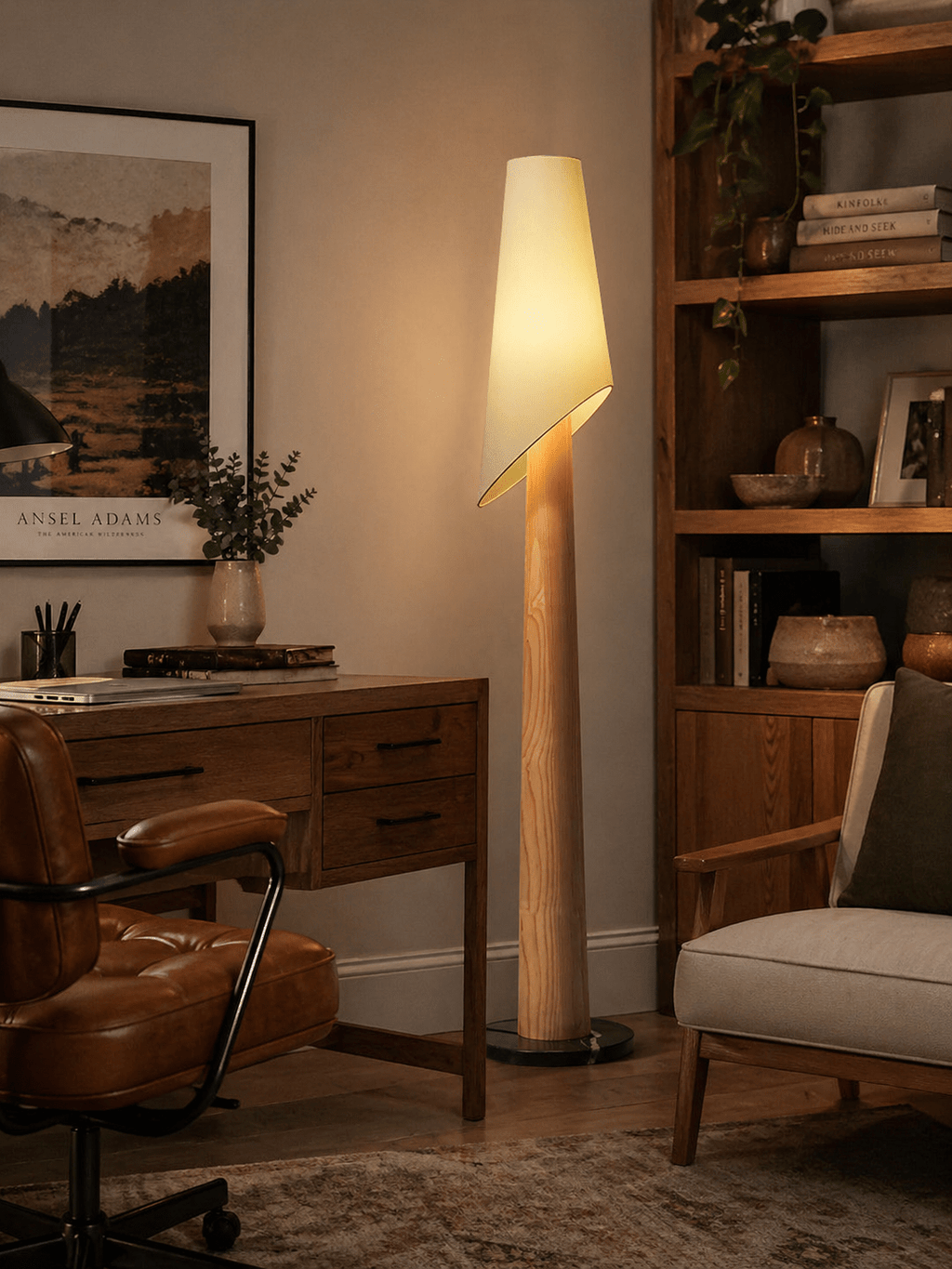 A Clérachic floor lamp lights a home office corner beside a wood desk, leather chair and bookshelf with plants.