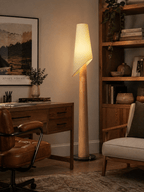 A Clérachic floor lamp lights a home office corner beside a wood desk, leather chair and bookshelf with plants.