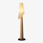 A Clérachic floor lamp stands lit against a plain grey backdrop, its angled fabric shade tilted over a wood-toned body.