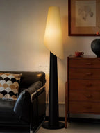 This black Clérachic floor lamp stands beside a leather chair and a wood dresser holding a small teacup.