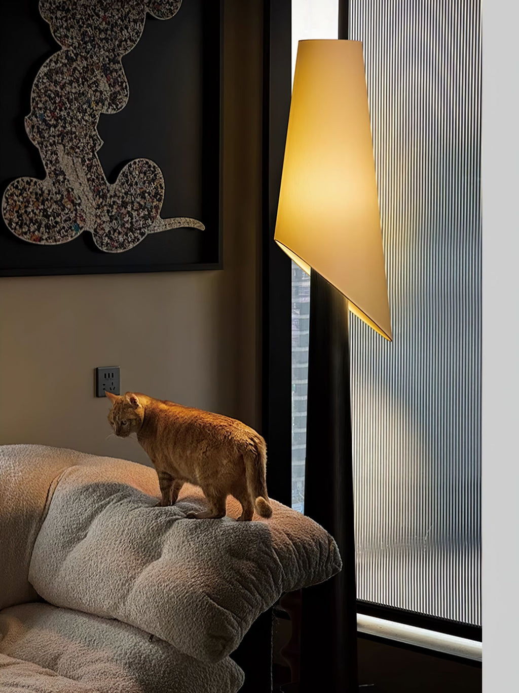 An orange cat perches on a cushioned chair beneath the lit shade of a dark Clérachic floor lamp.